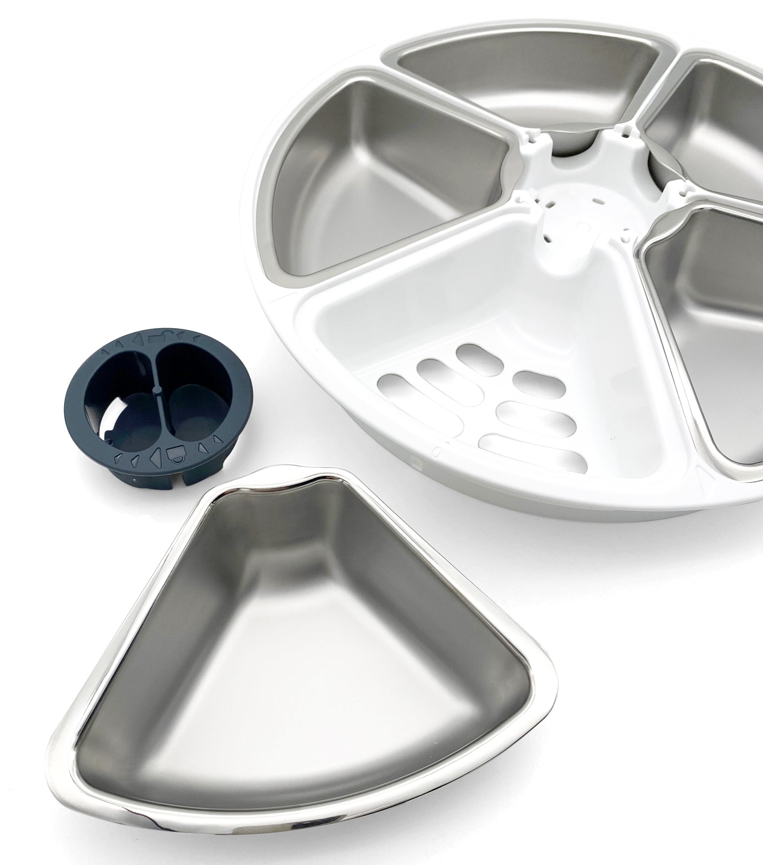 Disassembled parts of an automatic pet feeder, including a white circular rotating base with five triangular stainless steel food compartments, one of which is removed and placed in the foreground.